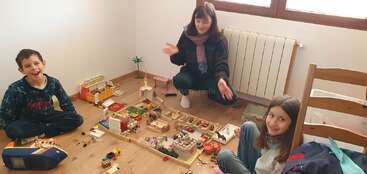 Das Bild zeigt eine Gruppe von Kindern, die mit Spielzeug spielen, mit einem Tisch und Stühlen im Hintergrund. Die Szene scheint ein unbeschwerter und fröhlicher Moment des kindlichen Spiels zu sein.