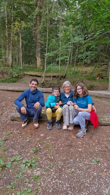 Vier Menschen und ein kleiner schwarzer Hund sitzen auf einem Baumstamm im Wald, lächeln und genießen einen friedlichen Moment, umgeben von hohen grünen Bäumen und der Natur.