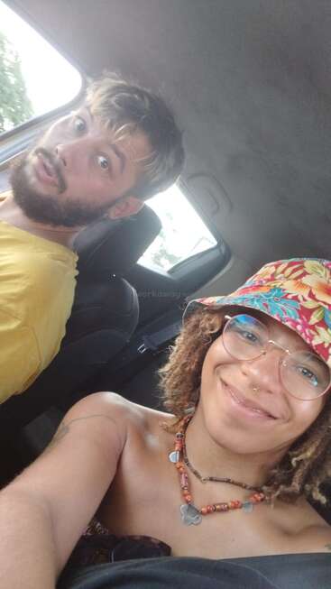 Two people are sitting in a car, smiling for a selfie. The woman wears a colorful hat and glasses; the man has a beard and yellow shirt.