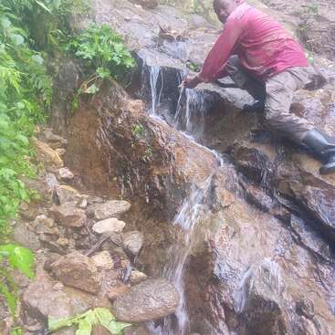 Um homem está agachado nas pedras ao lado de uma pequena cachoeira, com as mãos na água, vestindo camisa vermelha, calça cáqui e botas pretas.