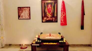 Esta imagen muestra un sencillo santuario casero con fotografías de un santo y una diosa, un altar decorado, flores, una lámpara y paños sagrados colgados de la pared.