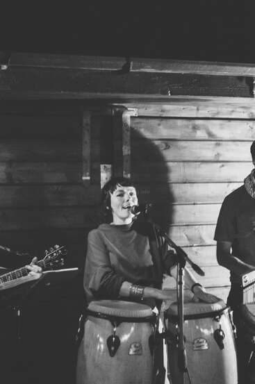 Una mujer toca la conga y canta con un micrófono. Está tocando con una banda, con un guitarrista visible, en un acogedor escenario interior de madera. En blanco y negro.