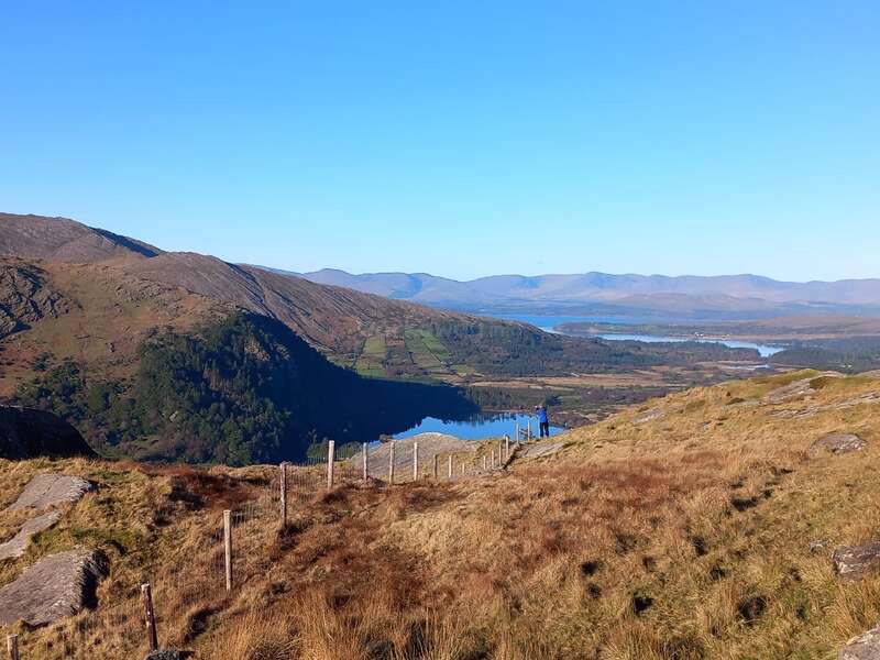 La imagen representa un paisaje sereno con colinas onduladas, un lago y montañas a lo lejos, bajo un cielo azul despejado en un día soleado.