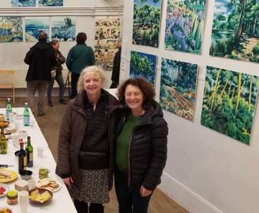 Two women stand smiling in an art gallery, surrounded by nature-inspired paintings. A long table displays snacks and drinks. Several people observe the artworks in the background.