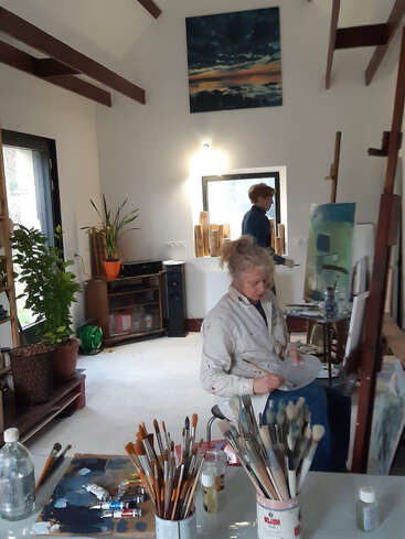 Dieses Bild zeigt zwei Personen, die in einem hellen, geräumigen Kunstatelier malen, mit vielen Pinseln, Pflanzen, Kunstzubehör auf dem Tisch und bunten Gemälden.