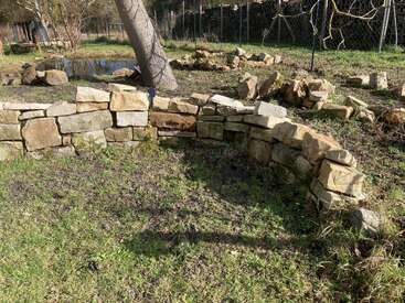 In einem Garten mit Gras steht eine geschwungene Steinmauer, die einen Baum teilweise umgibt. Im Hintergrund sieht man noch mehr Steine und einen kleinen Teich.