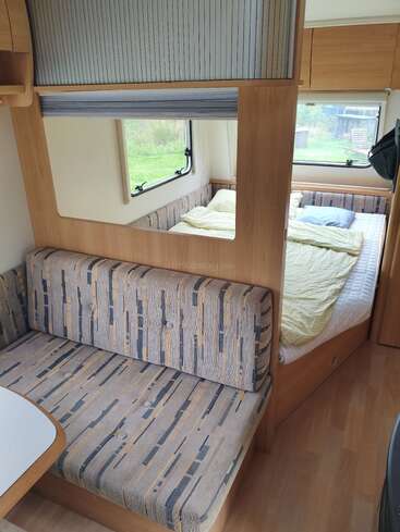 Esta imagen muestra el acogedor interior de una furgoneta camper, con una zona de asientos acolchada, una cama bien hecha, grandes ventanas y mobiliario con acentos de madera.
