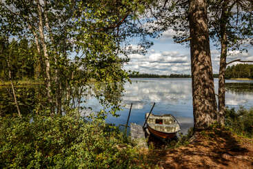 Una tranquila escena junto a un lago con una pequeña barca amarrada a un muelle de madera, rodeada de frondosos árboles verdes y aguas tranquilas que reflejan un cielo parcialmente nublado.