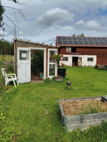 Una acogedora escena rural con un pequeño invernadero, una cama elevada de madera, una silla blanca, exuberante césped verde y un granero techado con paneles solares.