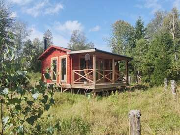 A charming red wooden cabin sits elevated in a grassy clearing, surrounded by tall green trees under a bright blue sky with scattered white clouds.