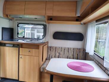 This image shows a cozy RV interior with wooden cabinets, a kitchenette, a small dining area, a bench seat, large windows, and a pink placemat.