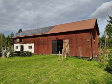 A large, red wooden barn with a sloped roof and solar panels sits on a grassy lawn, surrounded by trees under a partly cloudy sky. Peaceful rural setting.