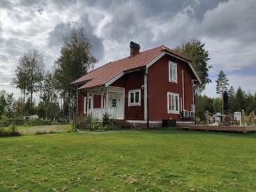 A charming red wooden house with a white porch sits amid lush green grass and tall trees under a cloudy sky, featuring an outdoor deck area.
