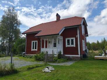 Uma charmosa casa de madeira vermelha com telhado de telha está sob um céu parcialmente nublado. Um cachorro descansa pacificamente no gramado verde da entrada.