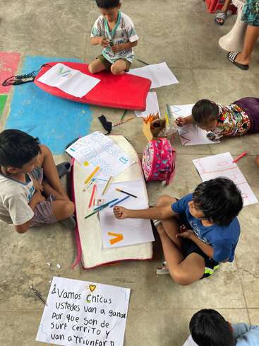 Several children are seated and lying on the floor, drawing and coloring on paper with markers and pencils. Handmade motivational posters in Spanish are visible among them.