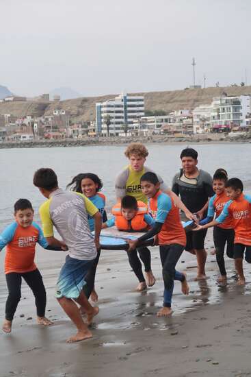 A group of smiling children and two adults, wearing wetsuits and colorful shirts, carry a boy on a surfboard along a sandy beach near the water.
