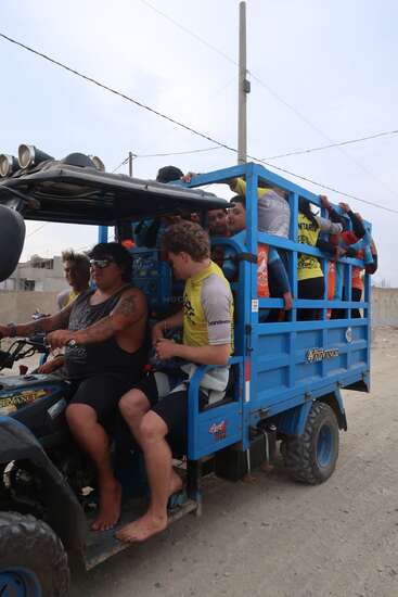 Eine Gruppe von Leuten, von denen einige Neoprenanzüge tragen, fährt gemeinsam in einem überfüllten blauen Geländewagen auf einer unbefestigten Straße und ist wahrscheinlich auf dem Weg zu einer Wassersportaktivität.