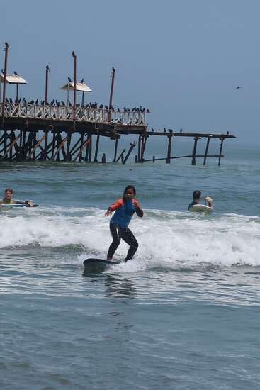 A young surfer rides a small wave near a wooden pier crowded with birds, while others paddle in the ocean under a clear blue sky.