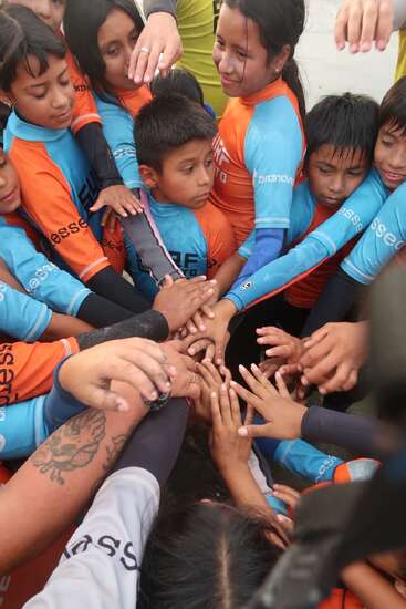 Eine Gruppe von Kindern in orangefarbenen und blauen T-Shirts versammelt sich im Kreis und legt die Hände ineinander – ein Symbol für Teamwork, Zusammenhalt, gegenseitige Unterstützung und Freundschaft.