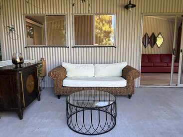Auf der gemütlichen Terrasse gibt's ein Korbsofa mit weißen Kissen, einen Couchtisch aus Metall mit Glasplatte, einen rustikalen Schrank und eine Schiebetür, hinter der man ein rotes Sofa sehen kann.