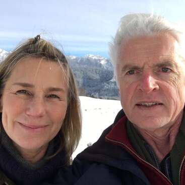 A man and woman take a selfie outdoors in a snowy mountain landscape, both smiling, bundled up in warm clothing under a bright, clear sky.