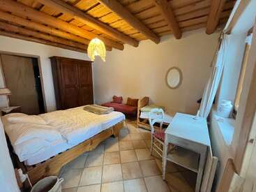 This cozy bedroom features a wooden-beam ceiling, double bed, armoire, daybed, desk with chair, lamp, mirror, tiled floor, and large window with natural light.