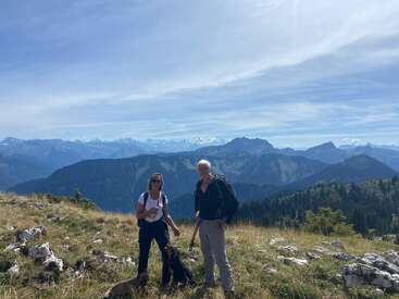 Two people and two dogs are standing on a grassy mountain slope, surrounded by rocks, with a stunning panoramic view of distant mountain ranges under a blue sky.