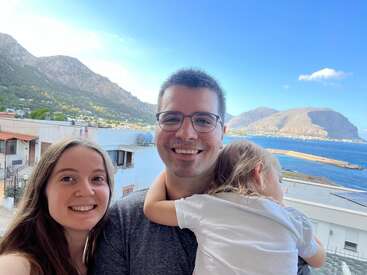 Das Bild zeigt eine dreiköpfige Familie, die für ein Selfie auf einem Balkon mit Blick auf ein ruhiges Gewässer und majestätische Berge unter einem klaren blauen Himmel posiert.