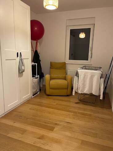 Ein gemütliches Zimmer mit Holzböden, einem gelben Sessel, einem Kleiderschrank, einem Wäscheständer mit Kleidung, einem Fenster, einem roten Gymnastikball, weißen Wänden und einer hängenden Deckenlampe.