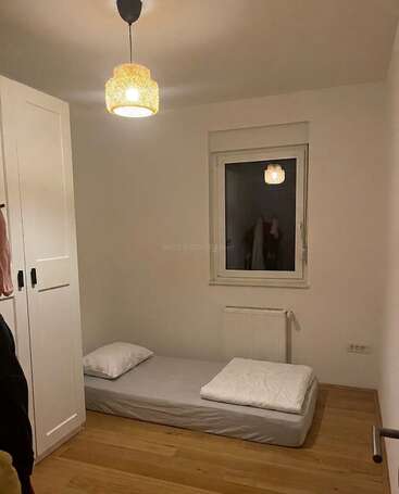 Ein minimalistisches Schlafzimmer mit Holzboden, weißen Wänden, einem Kleiderschrank, einer Einzelmatratze auf dem Boden mit Kissen und Decke sowie einer hängenden Deckenlampe.