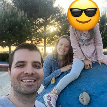 Une famille profite d'un moment en plein air dans un parc au coucher du soleil. L'homme sourit, la femme a l'air heureuse, et le visage de l'enfant est masqué par un emoji représentant des lunettes de soleil.