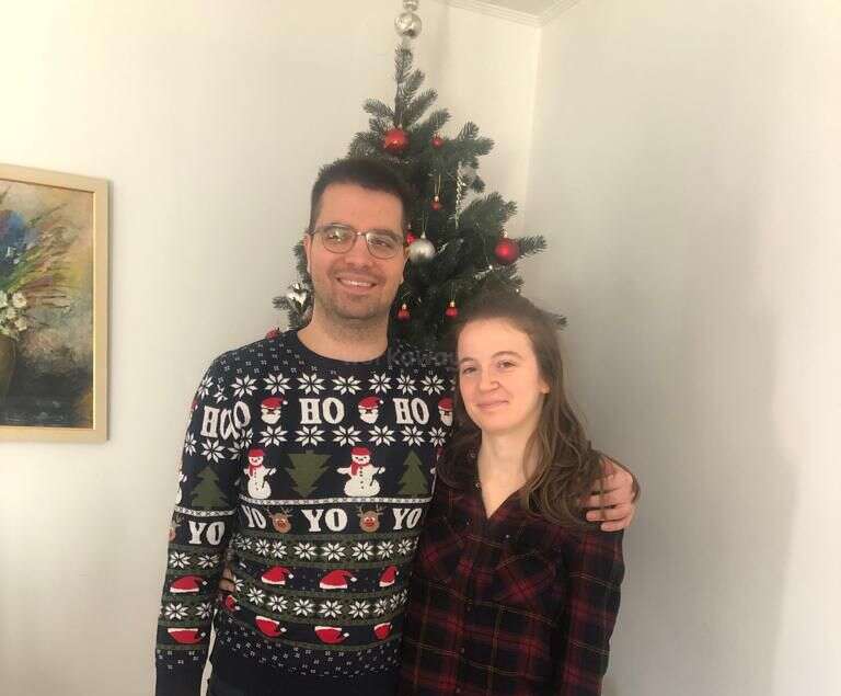 Das Bild zeigt einen Mann und eine Frau, die vor einem Weihnachtsbaum stehen, wobei der Mann einen festlichen Pullover und die Frau ein kariertes Hemd trägt.