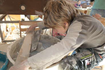 Un jeune enfant se concentre intensément sur son travail au tour de poterie, façonnant l'argile avec ses mains, dans un atelier désordonné mais créatif, éclairé par le soleil.