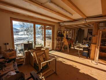 La lumière du soleil remplit un atelier de menuiserie confortable avec des poutres en bois, des outils et du bois d'œuvre éparpillés. De grandes fenêtres offrent une vue imprenable sur le paysage enneigé et les montagnes à l'extérieur.