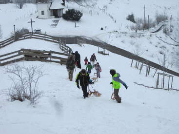 Des enfants et des adultes vêtus de vêtements d'hiver grimpent une colline enneigée en tirant des traîneaux en bois. Une petite église et des clôtures sont visibles à l'arrière-plan, créant une scène paisible.