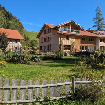 Uma charmosa casa de madeira com telhado vermelho fica em um jardim verde exuberante, cercada pela natureza, colinas, céu azul claro e uma cerca de madeira rústica.