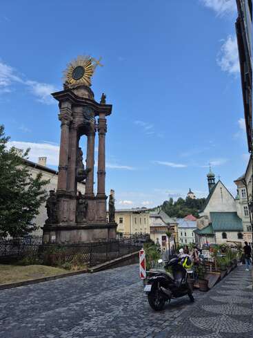 Un grand monument orné d'un soleil doré se dresse dans une rue pavée. À proximité, des personnes sont assises dans un café en plein air et un scooter est garé.