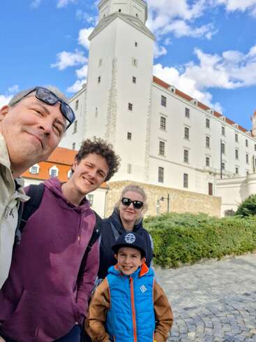 Eine fröhliche vierköpfige Gruppe lächelt vor einem großen, weißen historischen Gebäude mit Türmen, unter einem strahlend blauen Himmel, der mit flauschigen Wolken übersät ist.