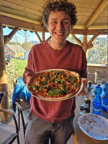 Ein lächelnder junger Mann in einem roten Pullover hält eine frisch gebackene Pizza mit Gemüse und Fleisch in der Hand und steht in einer gemütlichen Holzterrasse.