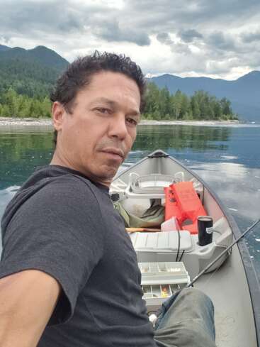 Ein Mann macht ein Selfie, während er in einem Kanu auf einem ruhigen See sitzt, umgeben von Bergen und Bäumen, mit Angelausrüstung und bedecktem Himmel.