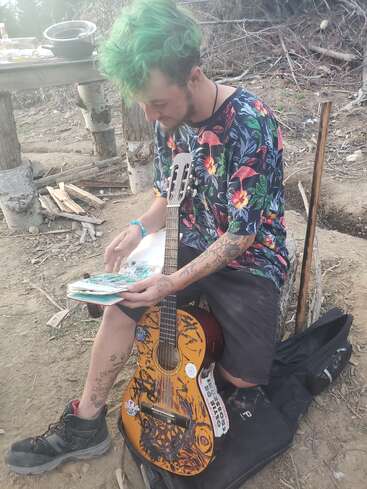 Um homem com cabelos verdes e tatuagens senta-se ao ar livre, vestindo uma camisa floral colorida. Ele lê, segurando um violão decorado, cercado por um cenário rústico e natural.