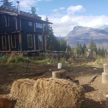 Una cabaña rústica en medio de un claro del bosque, con pacas de heno en primer plano y majestuosas montañas a lo lejos bajo un cielo parcialmente nublado.