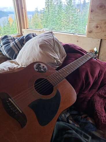 Scène de cabane confortable avec une guitare posée sur un lit recouvert de couvertures et d'oreillers, avec vue sur une belle forêt et un lac à travers de grandes fenêtres.
