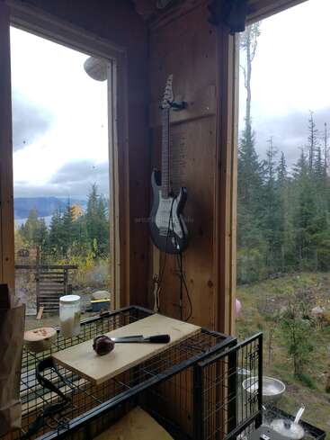 Une guitare électrique est accrochée à un mur en bois entre deux grandes fenêtres avec vue sur la forêt. En dessous, une table contient un maillet, une jarre et d'autres outils.