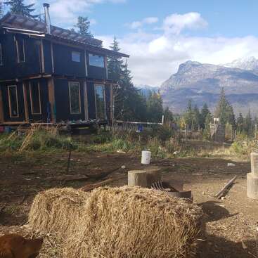 A rustic cabin sits amidst a forest clearing, with hay bales in the foreground and majestic mountains in the distance under a partly cloudy sky.