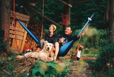 Two friends relax in a hammock with a golden dog, surrounded by nature, wood piles, a guitar, and greenery, enjoying a peaceful outdoor moment together.