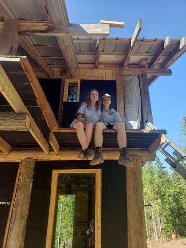 Two people sit together on the unfinished balcony of a wooden cabin, smiling under a clear blue sky, surrounded by forest, enjoying the construction progress.