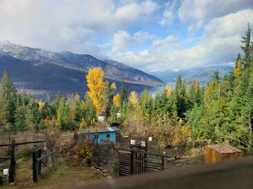 A scenic mountain landscape featuring green and yellow trees, a small blue house, wooden structures, and a distant river under a partly cloudy sky with sunlight.