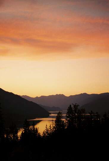 A imagem captura um pôr do sol sereno sobre um lago tranquilo, cercado por montanhas e árvores em silhueta, com um céu dourado refletindo na água calma abaixo.