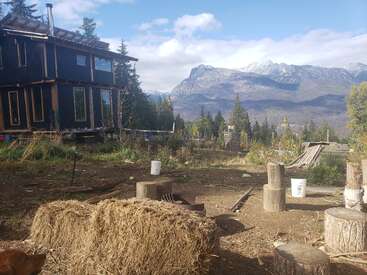 A rustic cabin sits amidst a forest clearing, with hay bales in the foreground and majestic mountains in the distance under a partly cloudy sky.